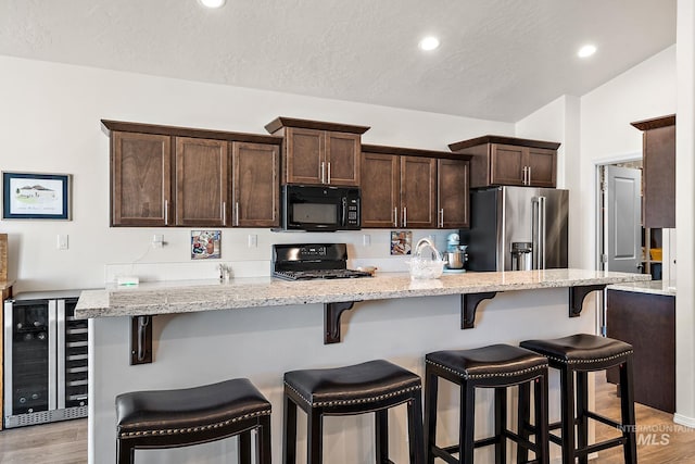 kitchen with wine cooler, a kitchen bar, dark brown cabinets, light wood-type flooring, and black appliances