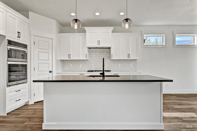 kitchen featuring dark countertops, wood finished floors, stainless steel double oven, white cabinetry, and a kitchen island with sink