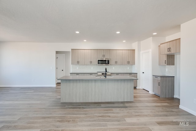 kitchen with light wood finished floors, stainless steel microwave, a sink, light brown cabinets, and a kitchen island with sink