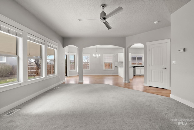 unfurnished living room featuring arched walkways, visible vents, light colored carpet, and ceiling fan with notable chandelier