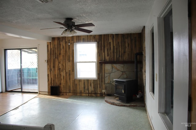 unfurnished living room featuring ceiling fan, a healthy amount of sunlight, a wood stove, and wooden walls