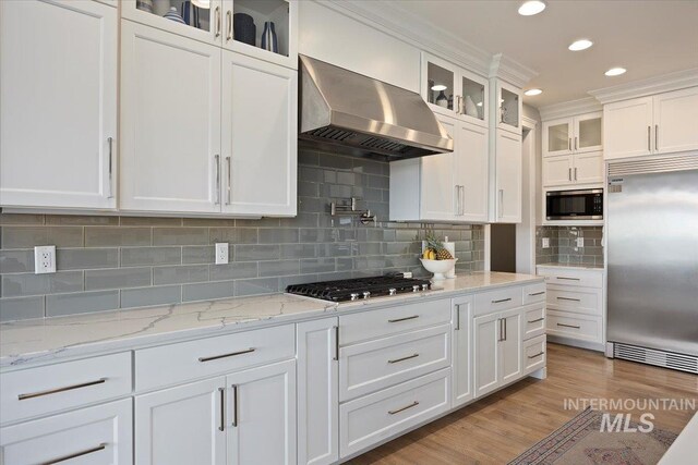 kitchen with tasteful backsplash, wall chimney range hood, built in appliances, light wood-type flooring, and white cabinetry