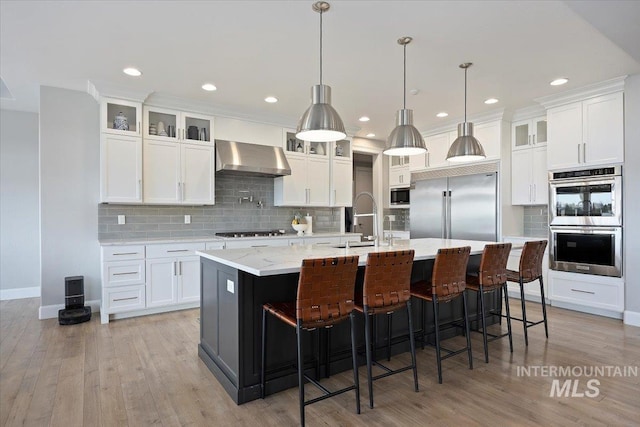 kitchen with a sink, light wood-style flooring, built in appliances, and wall chimney exhaust hood