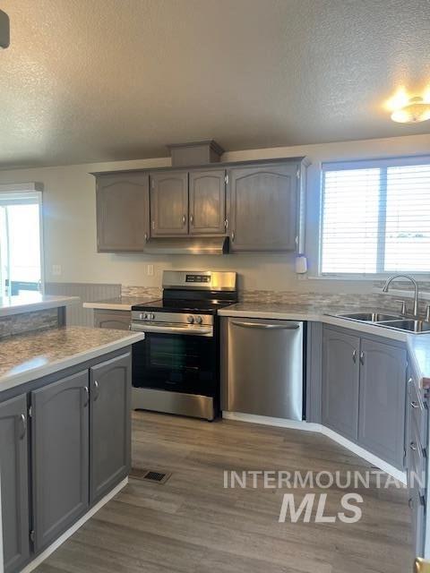 kitchen with a sink, under cabinet range hood, a textured ceiling, dark wood finished floors, and appliances with stainless steel finishes