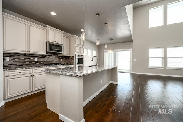 kitchen featuring a kitchen island with sink, stainless steel appliances, decorative light fixtures, light stone countertops, and backsplash