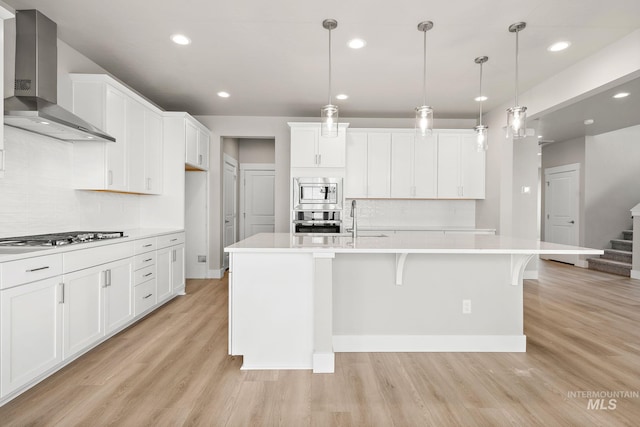 kitchen with stainless steel appliances, light countertops, white cabinetry, wall chimney exhaust hood, and light wood-type flooring