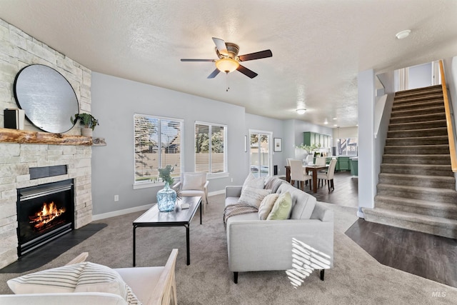 carpeted living room featuring a ceiling fan, a stone fireplace, a textured ceiling, baseboards, and stairs