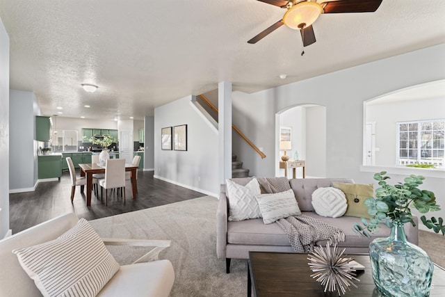 living room with baseboards, a ceiling fan, stairway, a textured ceiling, and dark carpet