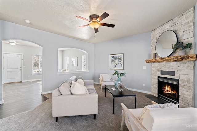 living room featuring arched walkways, a textured ceiling, a stone fireplace, a ceiling fan, and baseboards