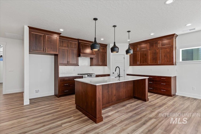 kitchen with pendant lighting, custom exhaust hood, light countertops, stove, and a kitchen island with sink