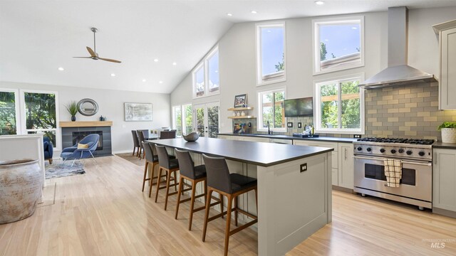 kitchen with high end stove, wall chimney range hood, dark countertops, light wood-type flooring, and high vaulted ceiling