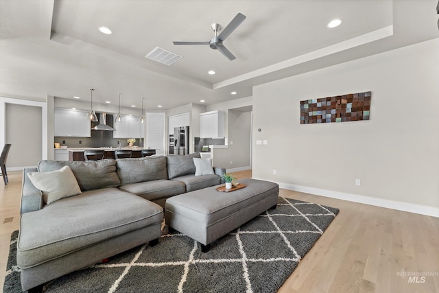 living area featuring a tray ceiling, a ceiling fan, light wood finished floors, baseboards, and recessed lighting