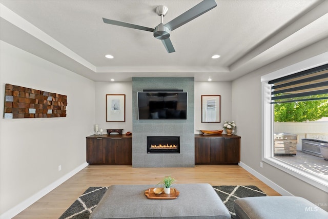 living area with a ceiling fan, a tiled fireplace, a tray ceiling, wood finished floors, and recessed lighting