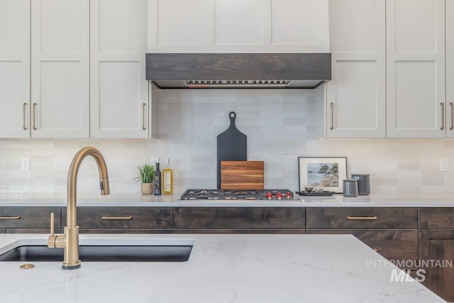 kitchen featuring a sink, decorative backsplash, light stone countertops, dark brown cabinetry, and white cabinetry