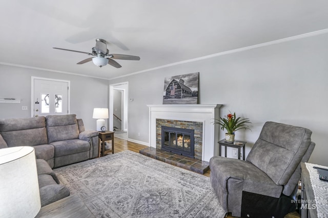 living area featuring wood finished floors, a stone fireplace, crown molding, baseboards, and ceiling fan