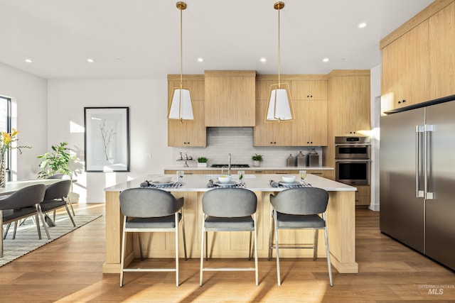 kitchen with stainless steel appliances, a kitchen island with sink, light brown cabinets, decorative light fixtures, and light hardwood / wood-style floors