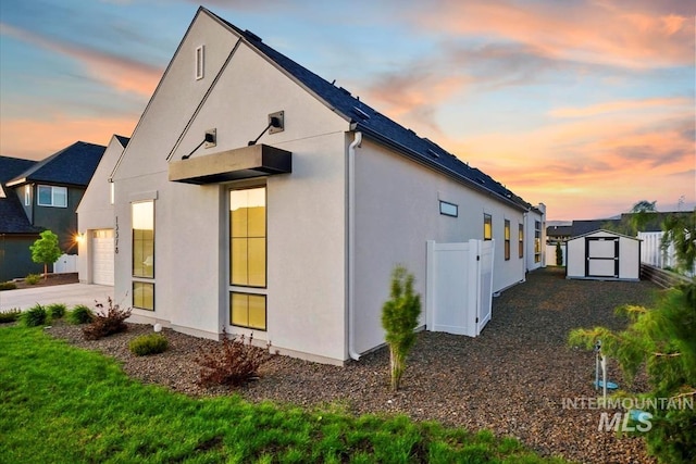 property exterior at dusk with a shed, stucco siding, fence, an outbuilding, and driveway