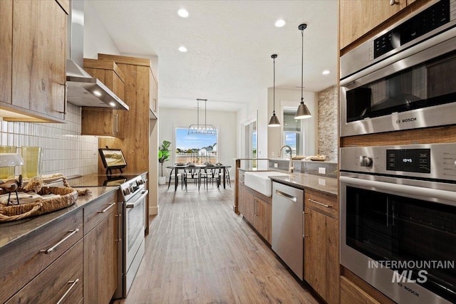 kitchen with light hardwood / wood-style floors, wall chimney exhaust hood, stainless steel appliances, decorative light fixtures, and sink