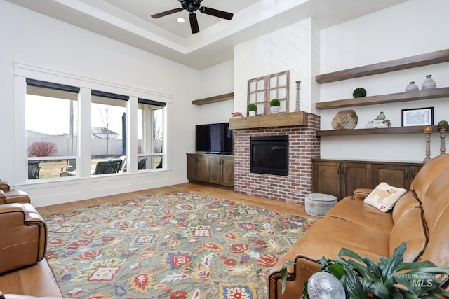 living room featuring a raised ceiling, light wood-style floors, a brick fireplace, and ceiling fan