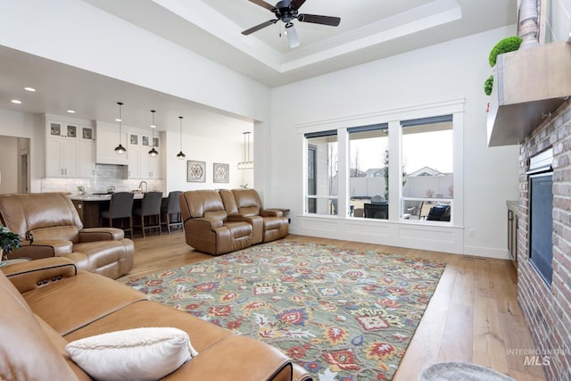 living area featuring a tray ceiling, a glass covered fireplace, recessed lighting, light wood-style floors, and ceiling fan