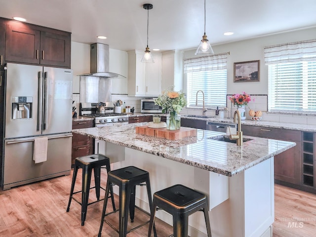 kitchen featuring wall chimney exhaust hood, appliances with stainless steel finishes, decorative light fixtures, a kitchen island, and light wood-type flooring