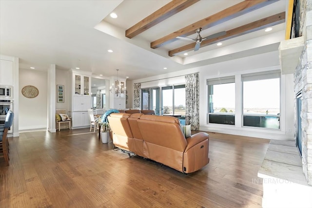 living area featuring beam ceiling, dark wood-type flooring, ceiling fan with notable chandelier, recessed lighting, and a stone fireplace