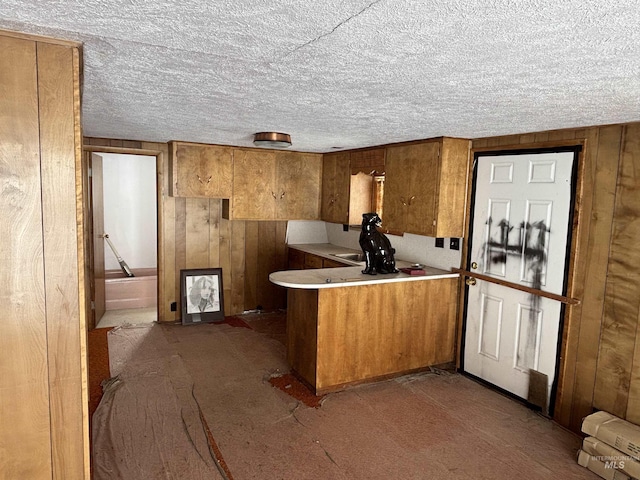 kitchen featuring a textured ceiling, a peninsula, wood walls, brown cabinetry, and light countertops