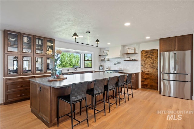 kitchen featuring glass insert cabinets, a center island, freestanding refrigerator, custom exhaust hood, and light wood-type flooring