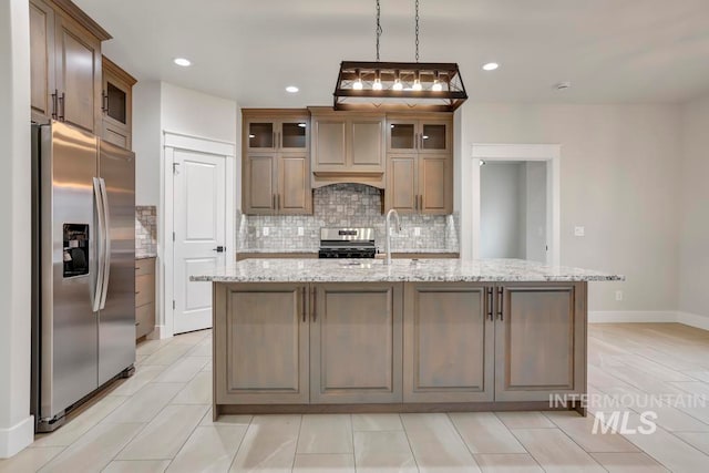 kitchen with light stone counters, glass insert cabinets, a sink, appliances with stainless steel finishes, and a center island with sink