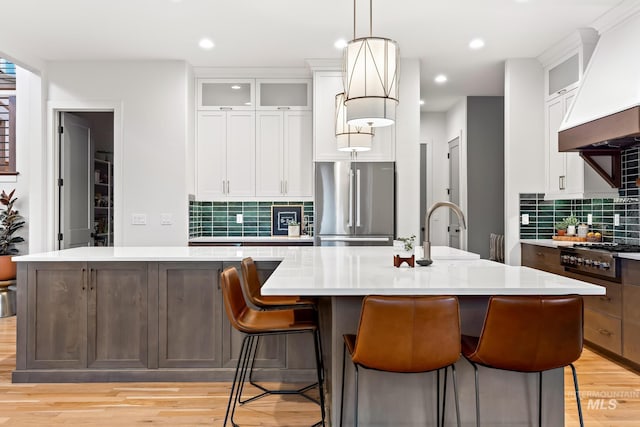 kitchen featuring appliances with stainless steel finishes, a kitchen island with sink, light countertops, premium range hood, and a sink