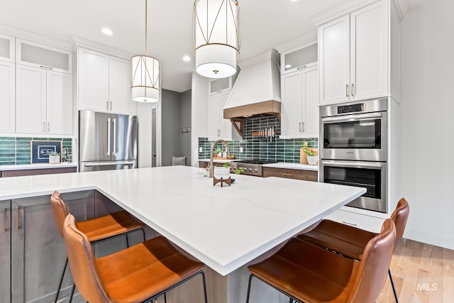 kitchen featuring stainless steel appliances, custom range hood, light wood finished floors, a kitchen bar, and tasteful backsplash