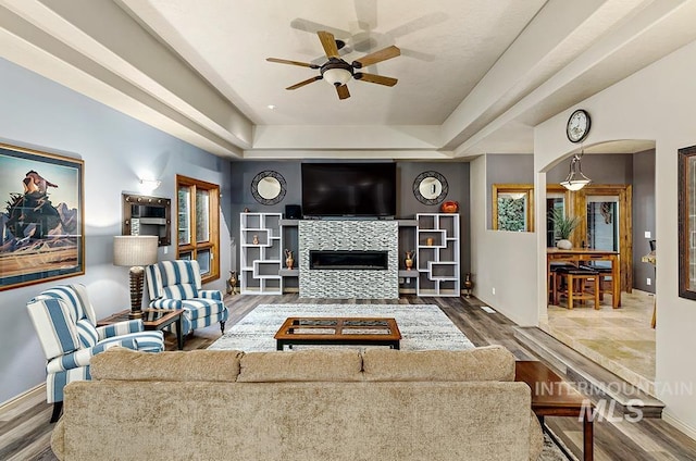 living room with baseboards, a glass covered fireplace, a ceiling fan, wood finished floors, and a raised ceiling