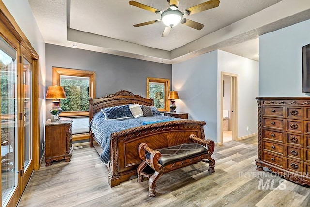 bedroom with ceiling fan, a tray ceiling, light wood finished floors, baseboards, and ensuite bath