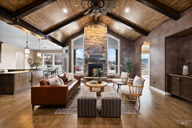 living area featuring arched walkways, a stone fireplace, ceiling fan with notable chandelier, wood finished floors, and beam ceiling