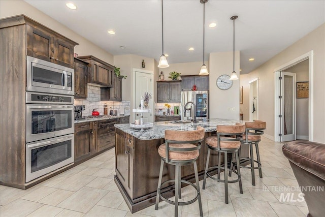4595 N Tirso Ave, Meridian, ID 83646 - kitchen with dark brown cabinetry, appliances with stainless steel finishes, light stone counters, an island with sink, and decorative backsplash