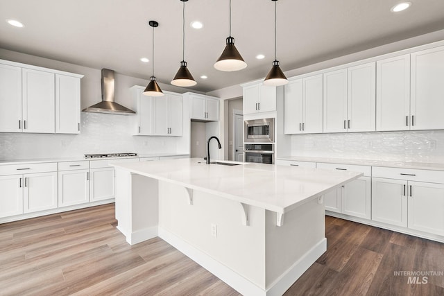 kitchen with wall chimney range hood, light wood-type flooring, stainless steel appliances, white cabinetry, and a sink