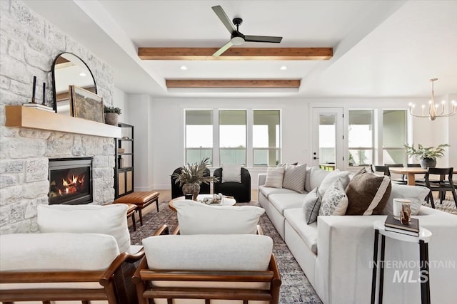 living room with wood finished floors, beam ceiling, a stone fireplace, and ceiling fan with notable chandelier