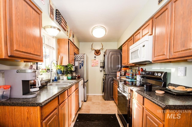 kitchen with stainless steel appliances, dark countertops, brown cabinetry, a sink, and stacked washing maching and dryer