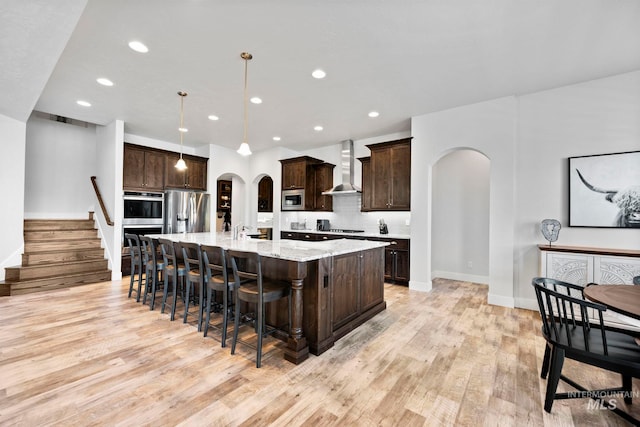 kitchen featuring arched walkways, dark brown cabinetry, appliances with stainless steel finishes, and wall chimney exhaust hood