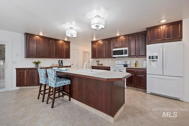 kitchen featuring light countertops, white appliances, a center island with sink, and dark brown cabinetry