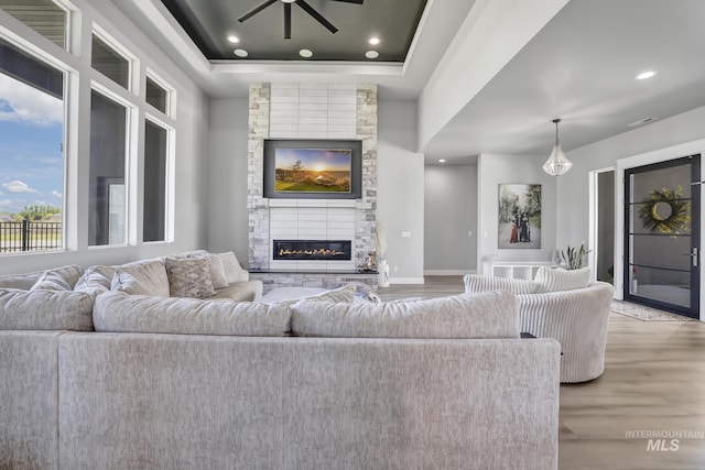 living room featuring ceiling fan, a stone fireplace, a raised ceiling, and light wood-style flooring