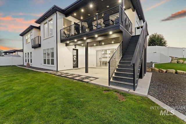 back house at dusk featuring a balcony, a lawn, and a patio area