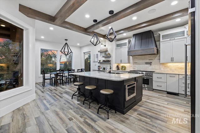 kitchen featuring custom exhaust hood, pendant lighting, a center island with sink, high end stainless steel range oven, and white cabinets