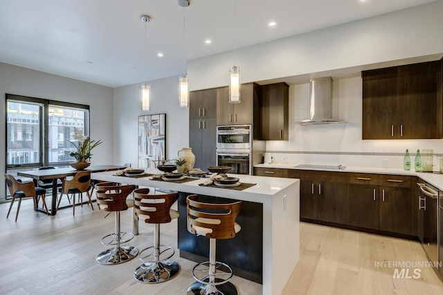 kitchen with wall chimney exhaust hood, decorative light fixtures, a center island, appliances with stainless steel finishes, and light hardwood / wood-style floors