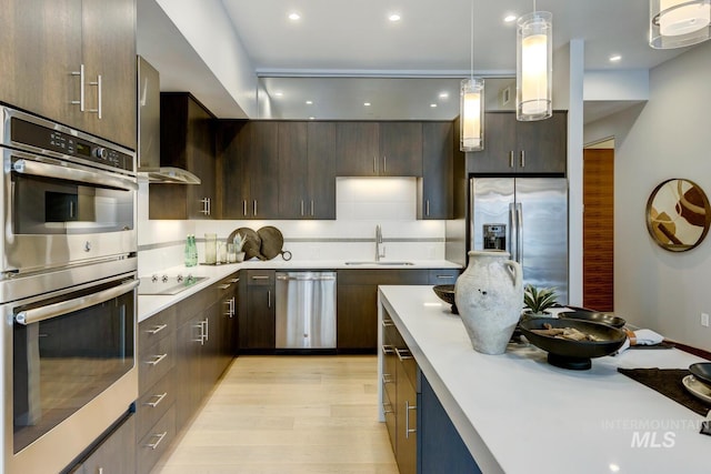 kitchen with wall chimney exhaust hood, dark brown cabinetry, sink, decorative light fixtures, and appliances with stainless steel finishes