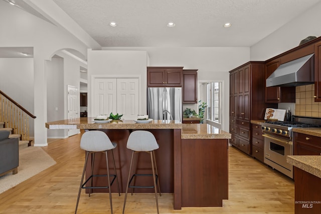 kitchen featuring appliances with stainless steel finishes, a kitchen island with sink, wall chimney range hood, and light hardwood / wood-style flooring