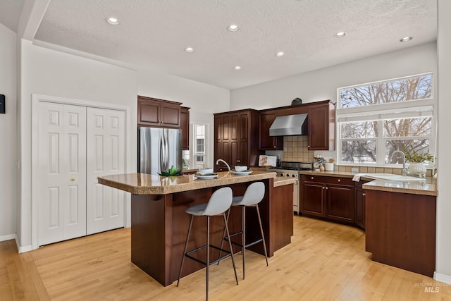 kitchen with wall chimney exhaust hood, a kitchen bar, sink, appliances with stainless steel finishes, and an island with sink