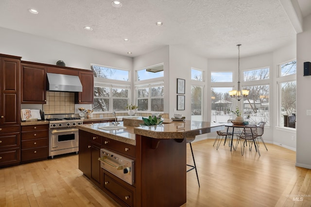 kitchen with wall chimney exhaust hood, high end range, a kitchen breakfast bar, an island with sink, and pendant lighting