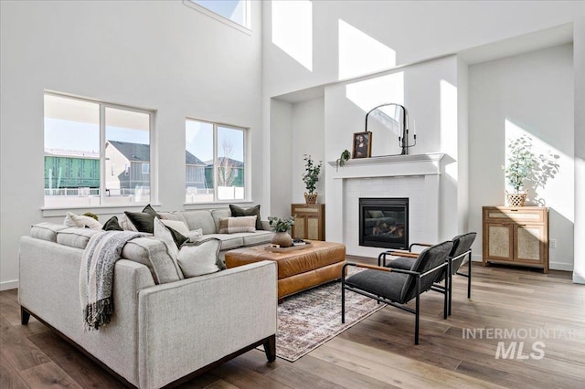 living room with a brick fireplace, wood finished floors, baseboards, and a towering ceiling