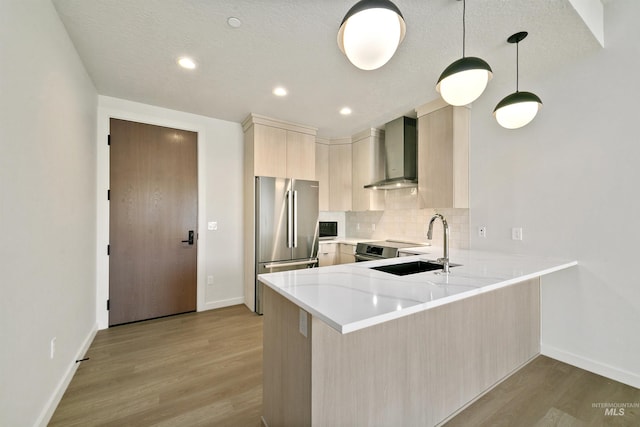 kitchen with a peninsula, wall chimney exhaust hood, a sink, light wood-style flooring, and stainless steel appliances